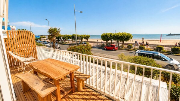 A balcony with a wooden table and chairs, overlooking a beach with cars and people.
