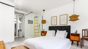 Ein Schlafzimmer mit einem Bett, einem Nachttisch aus Holz, einem Sessel, einer Leiter und einer Tür, die auf einen Balkon führt.