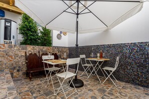 A patio area with a table and chairs, a white umbrella, a mosaic tile wall, and a water heater. - Tavira Bliss Garden Apartment (Tavira)