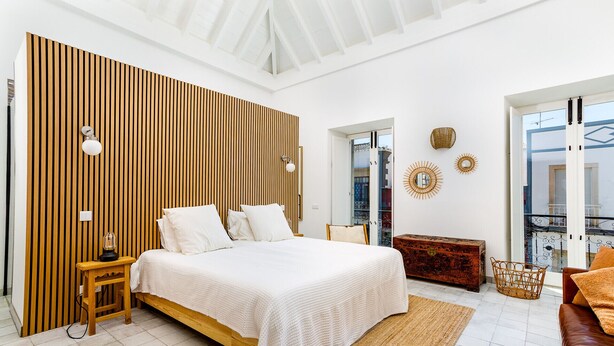 Ein modernes Schlafzimmer mit einer Wand aus Holzlatten, einem Bett mit weißen Leinen, einem Nachttisch aus Holz, einem Korbstuhl und Blick ins Freie.