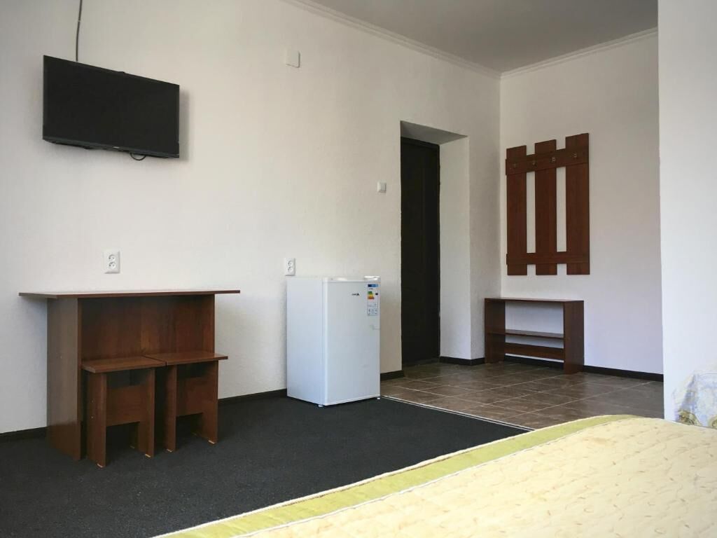 Una habitación con una cama, un escritorio pequeño, un refrigerador blanco, un televisor montado en la pared y una puerta que da a otra habitación.