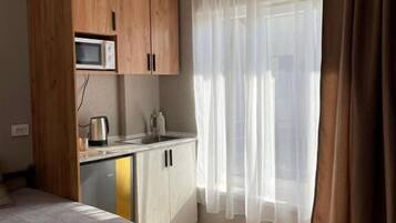 A room with a bed, a kitchenette, a microwave, a kettle, and a window with curtains.