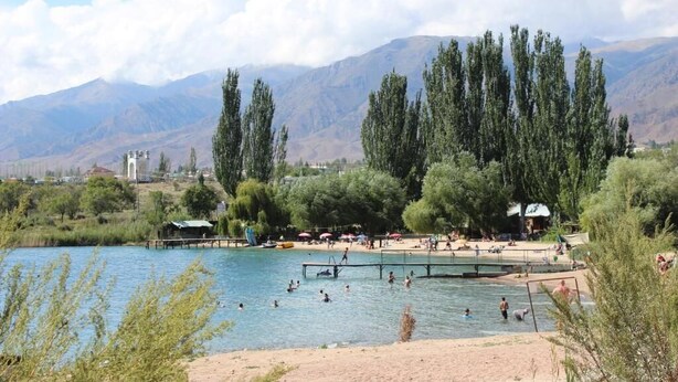 A lakeside swimming area with a dock, surrounded by trees and mountains in the background.