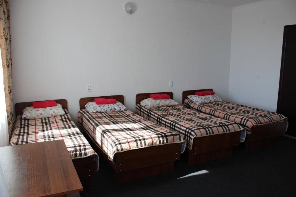 Une chambre avec quatre lits simples, chacun recouvert d’une couverture à carreaux et agrémenté d’un oreiller rouge. Une table en bois se trouve au centre de la pièce.