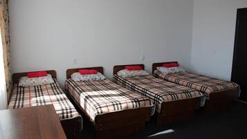 Une chambre avec quatre lits simples, chacun recouvert d’une couverture à carreaux et agrémenté d’un oreiller rouge. Une table en bois se trouve au centre de la pièce.