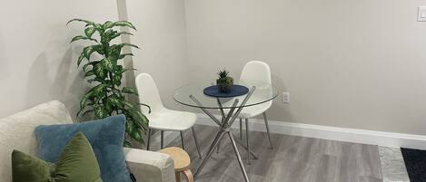 A living room with a glass-top table, two white chairs, a white sofa with green and blue cushions, and a large green plant.