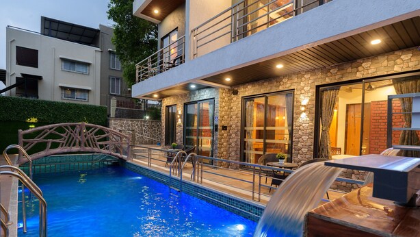 A pool with a waterfall feature, a stone facade, and a balcony overlooking the pool. - Sunflower Vista - 6BR Estate with Pool, Hot Tub & Hill Views, Mahabaleshwar (Panchgani)