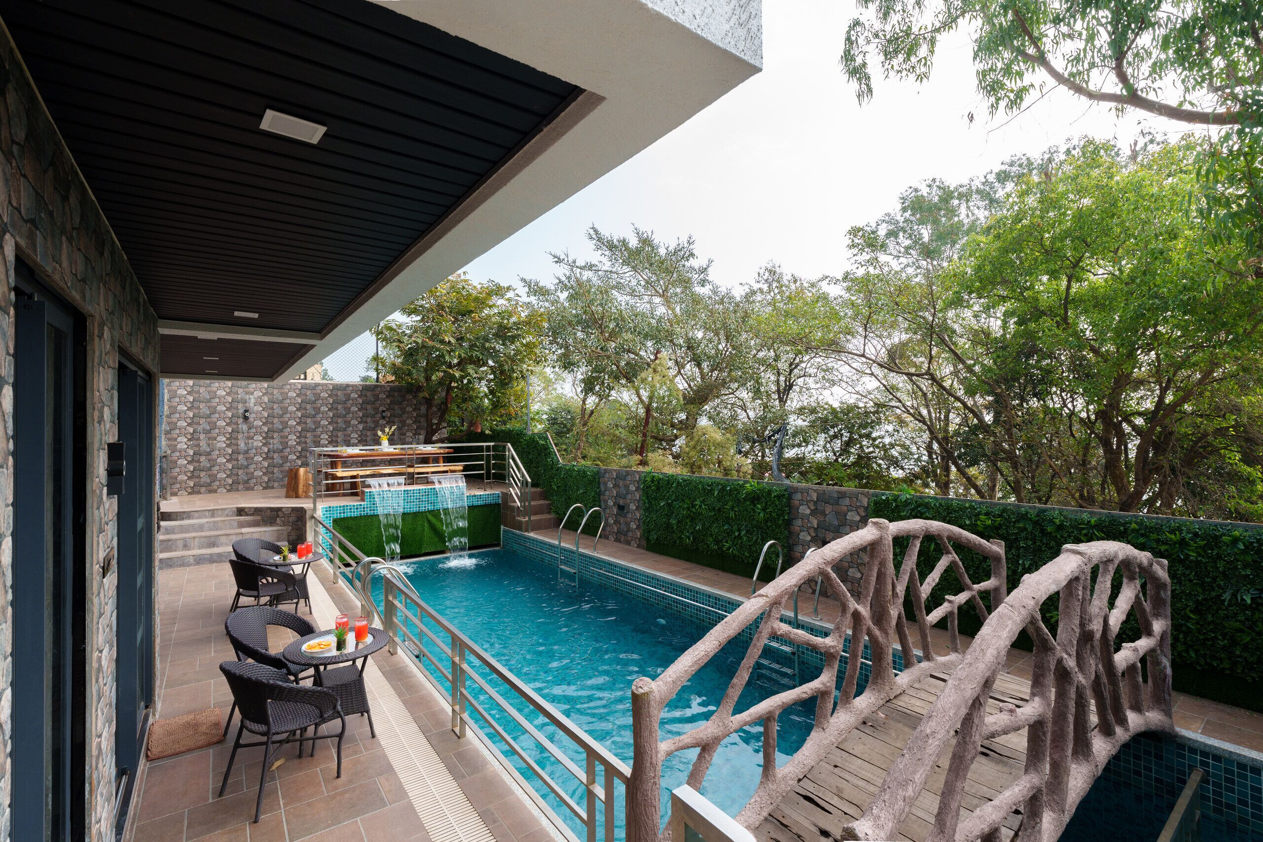 An outdoor swimming pool with a wooden bridge, stone walls, and a patio area with chairs and tables.
