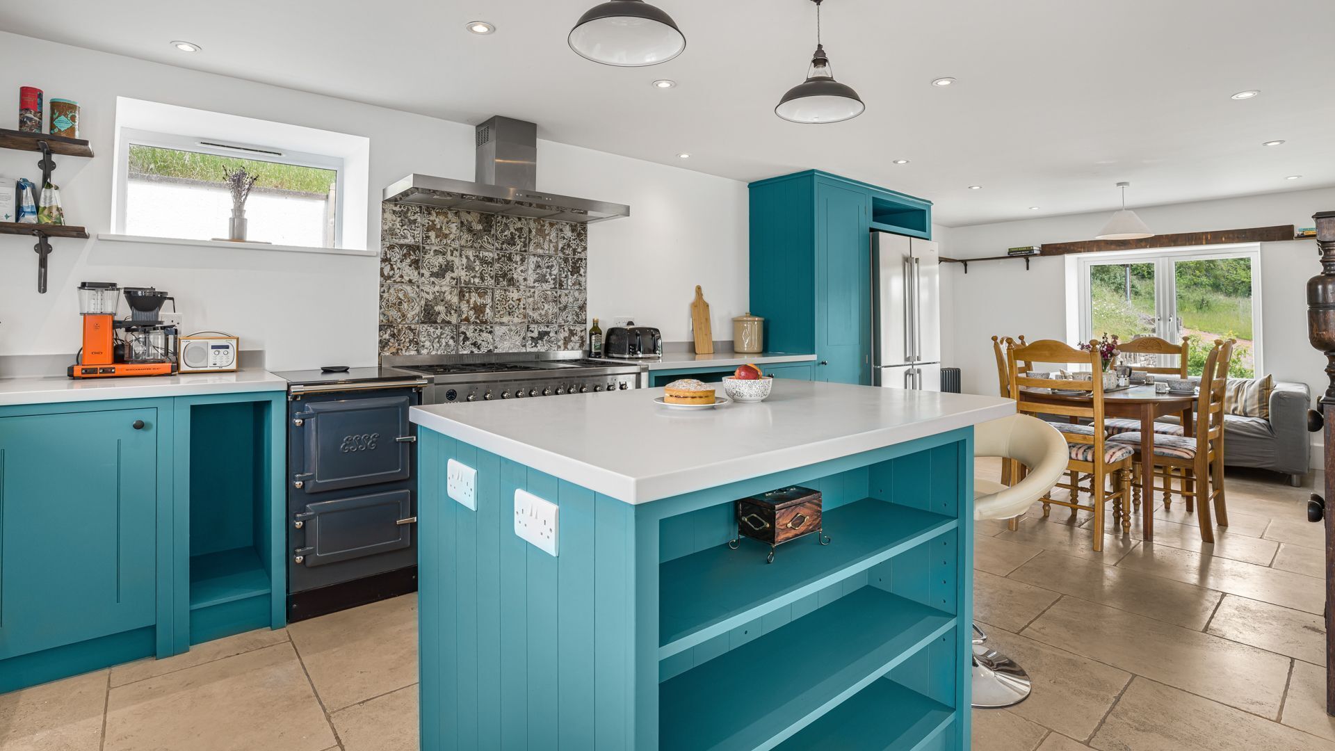 A modern kitchen with teal cabinetry, a central island, and a dining area with a wooden table and chairs.