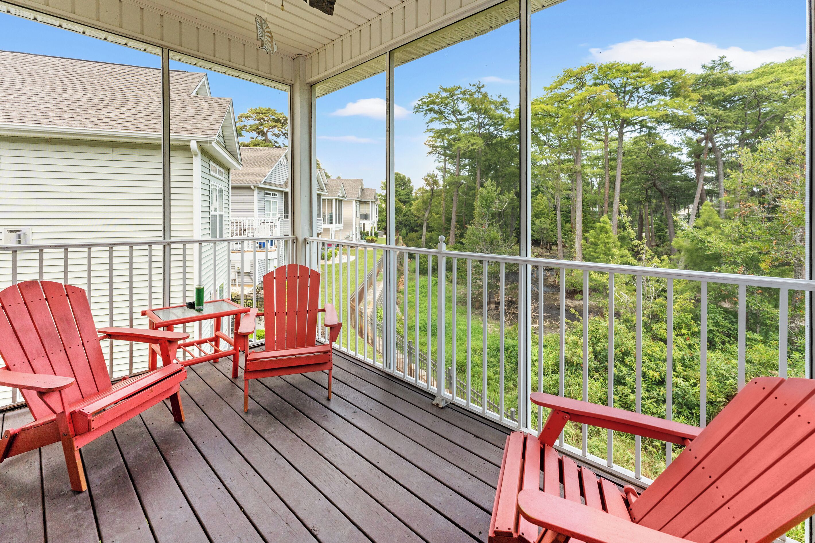 A covered porch with red Adirondack chairs and a table, overlooking a residential area with trees and houses.