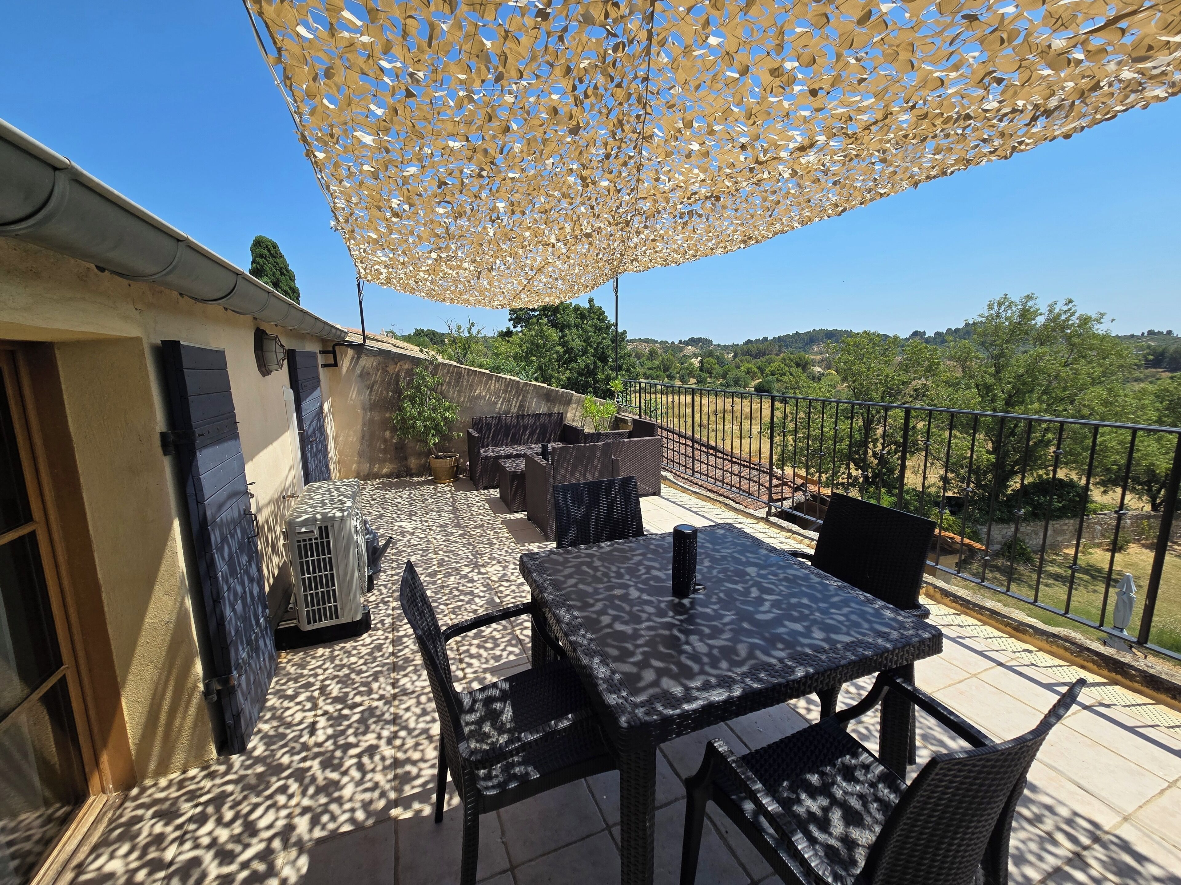 A balcony with outdoor furniture, a table, and chairs, overlooking a landscape with trees and a fence.