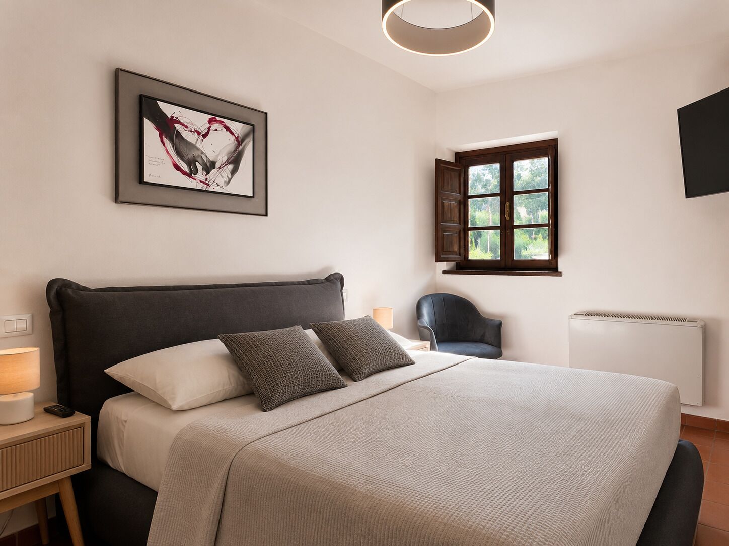 Ein Schlafzimmer mit einem Bett, einem Sessel, einem Fenster, einer Heizung und einem an der Wand hängenden Kunstwerk.