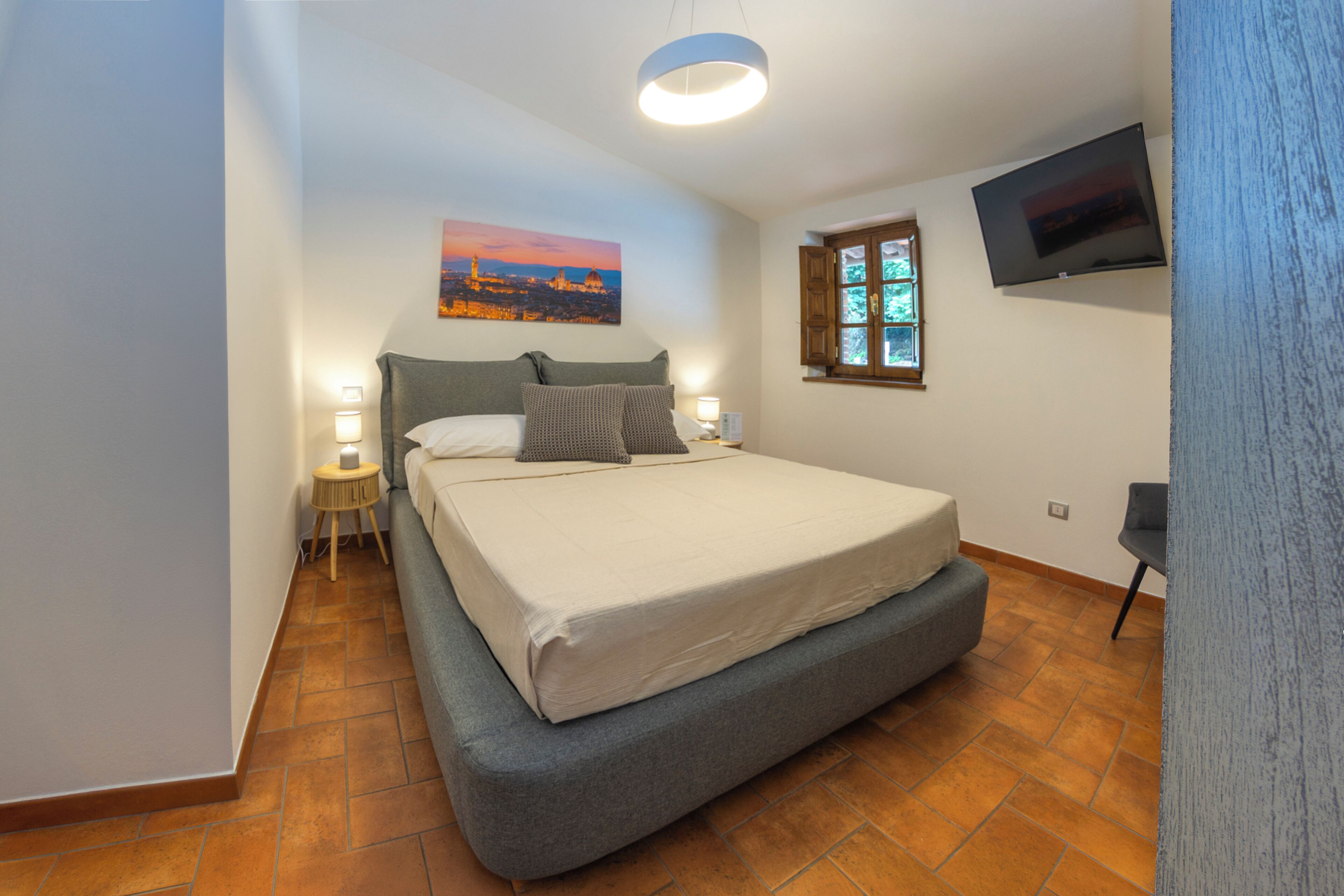 Ein Schlafzimmer mit einem Bett, einem Nachttisch, einer Lampe, einem Gemälde, einem Fenster und einem Fernseher.