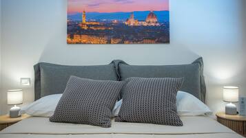 Un letto rifatto con due cuscini decorati, un quadro con una veduta cittadina sopra e due lampade ai lati.