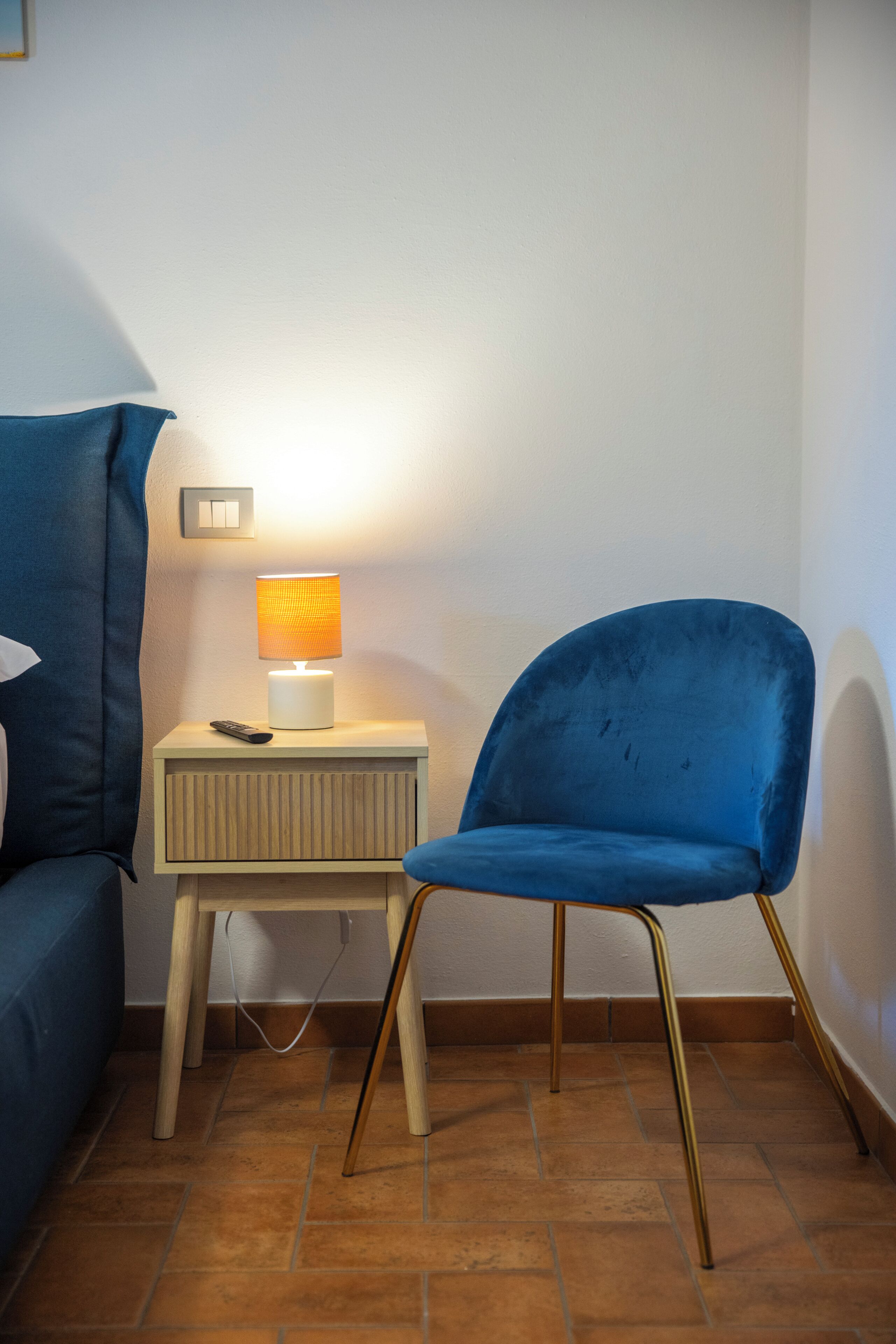 A corner of a room with a blue chair, a small bedside table with a lamp, and a power outlet.