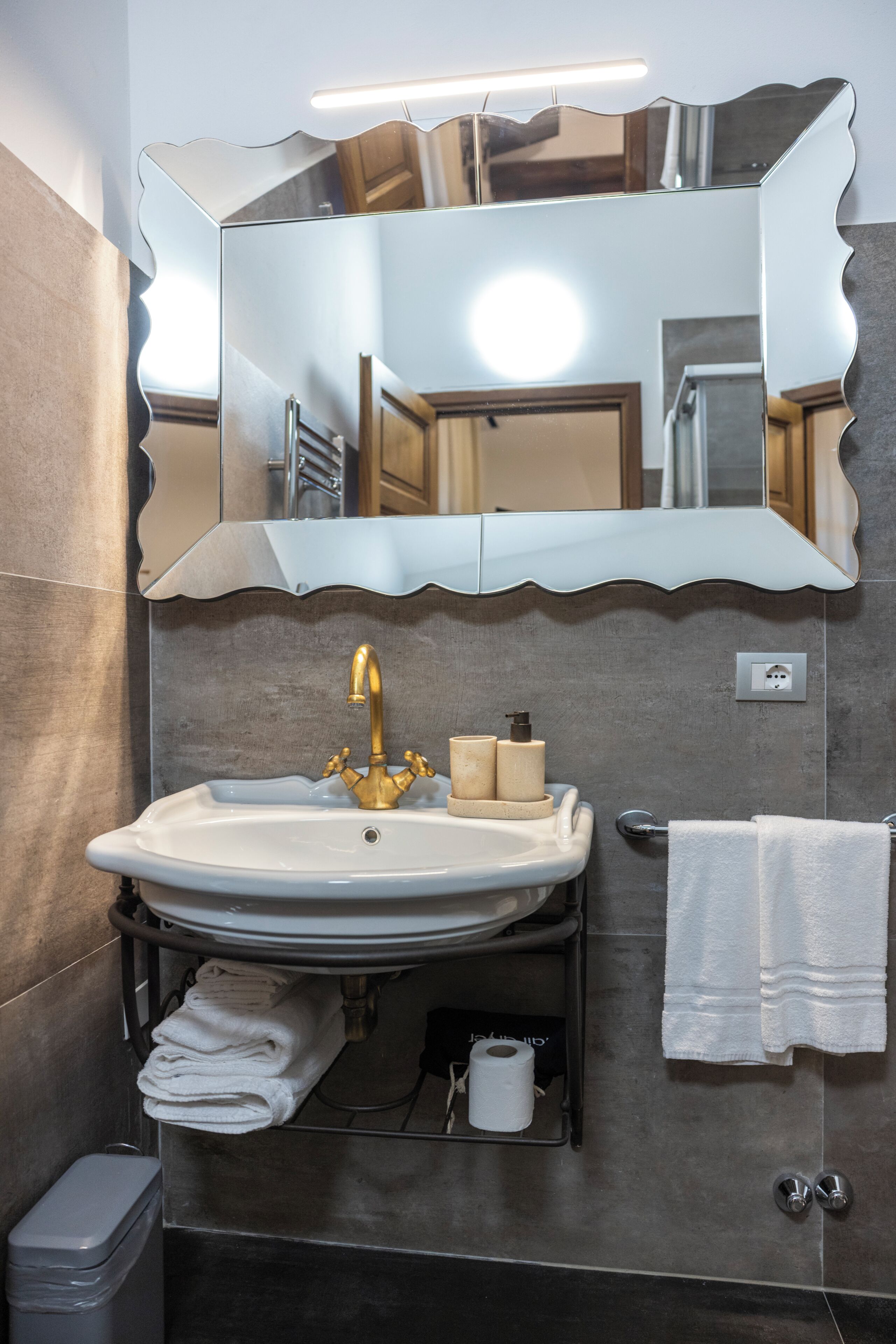 Un baño con lavamanos, espejo, perchero y cubo de basura.
