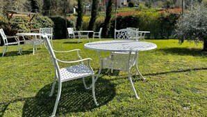 A garden with a round metal table and white metal chairs, surrounded by greenery and trees. - Mulino La Sosta (Camaiore)