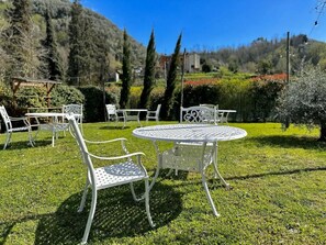 A garden with a round metal table and white metal chairs, surrounded by greenery and trees. - Mulino La Sosta (Camaiore)