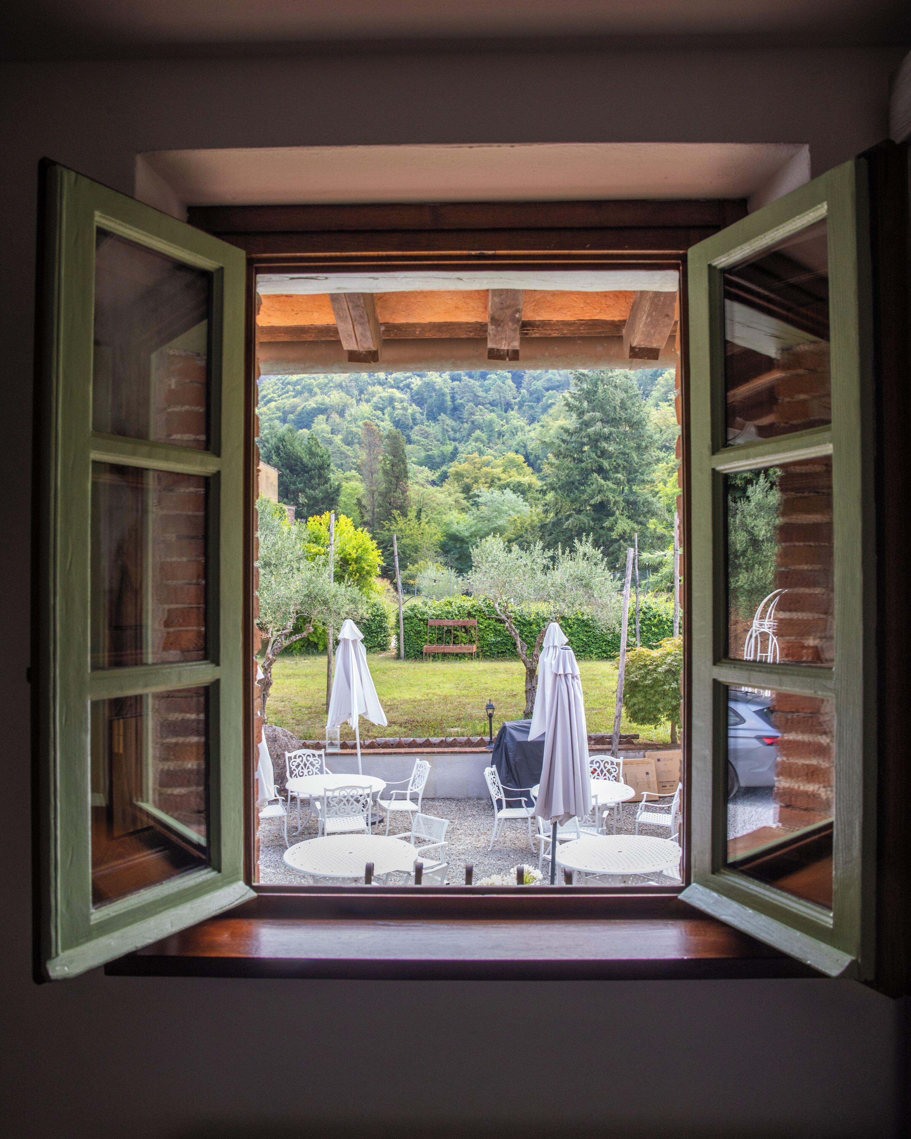 Ein offenes Fenster mit Blick auf eine Außenpatio-Terrasse, auf der Tisch und Stühle, ein Sonnenschirm und eine üppige grüne Landschaft zu sehen sind.
