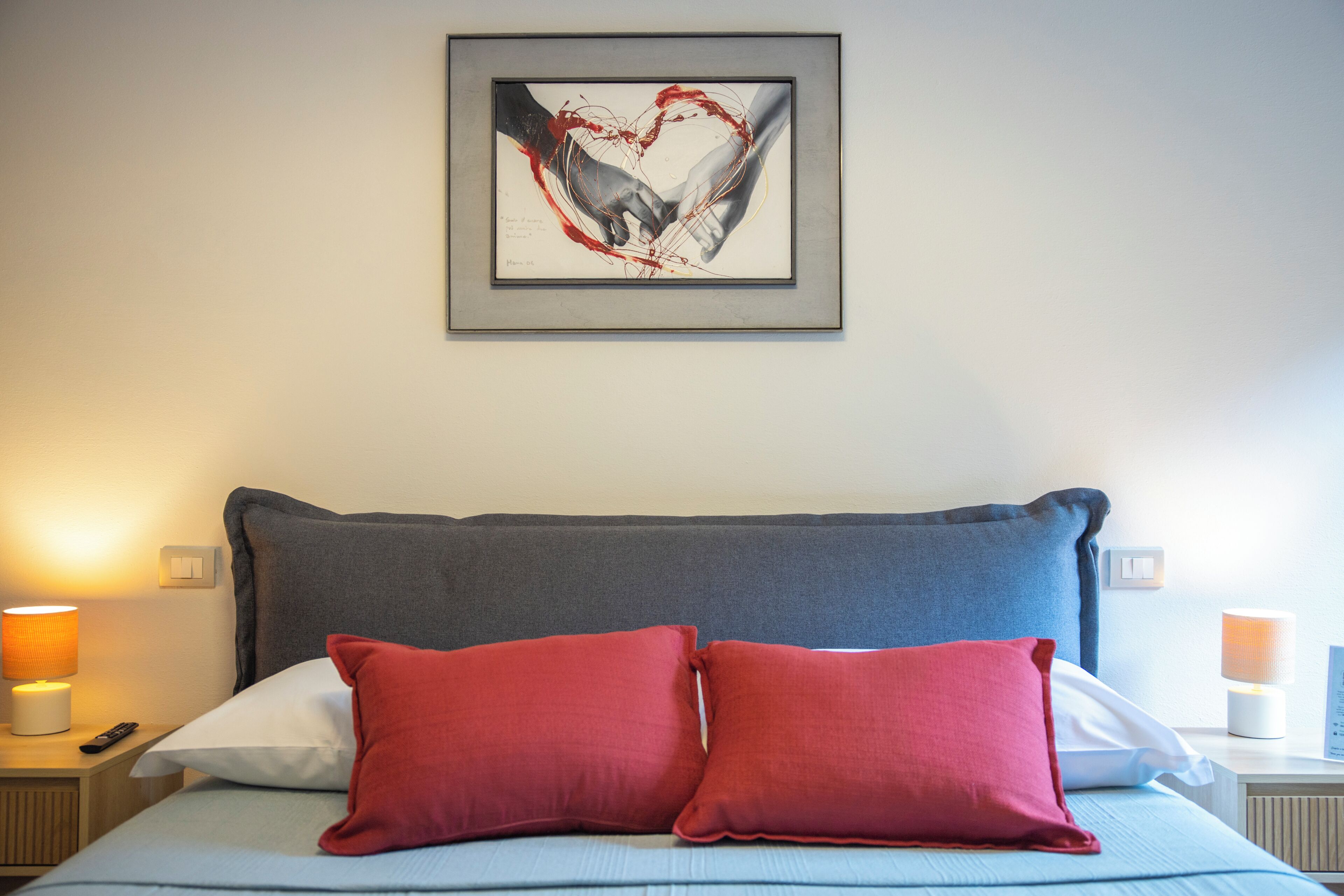Un dormitorio con una cama, dos almohadas rojas, una obra de arte enmarcada sobre la cama y lámparas de noche.