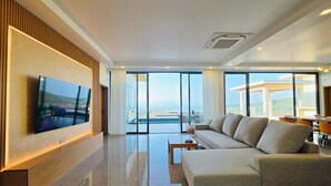 Een moderne woonkamer met een grote flatscreen televisie, een bank, een salontafel en uitzicht op zee door grote ramen.