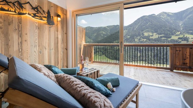 Una stanza con parete in legno, un balcone vista montagna, un letto con cuscini e un tavolo con bottiglia e bicchiere.