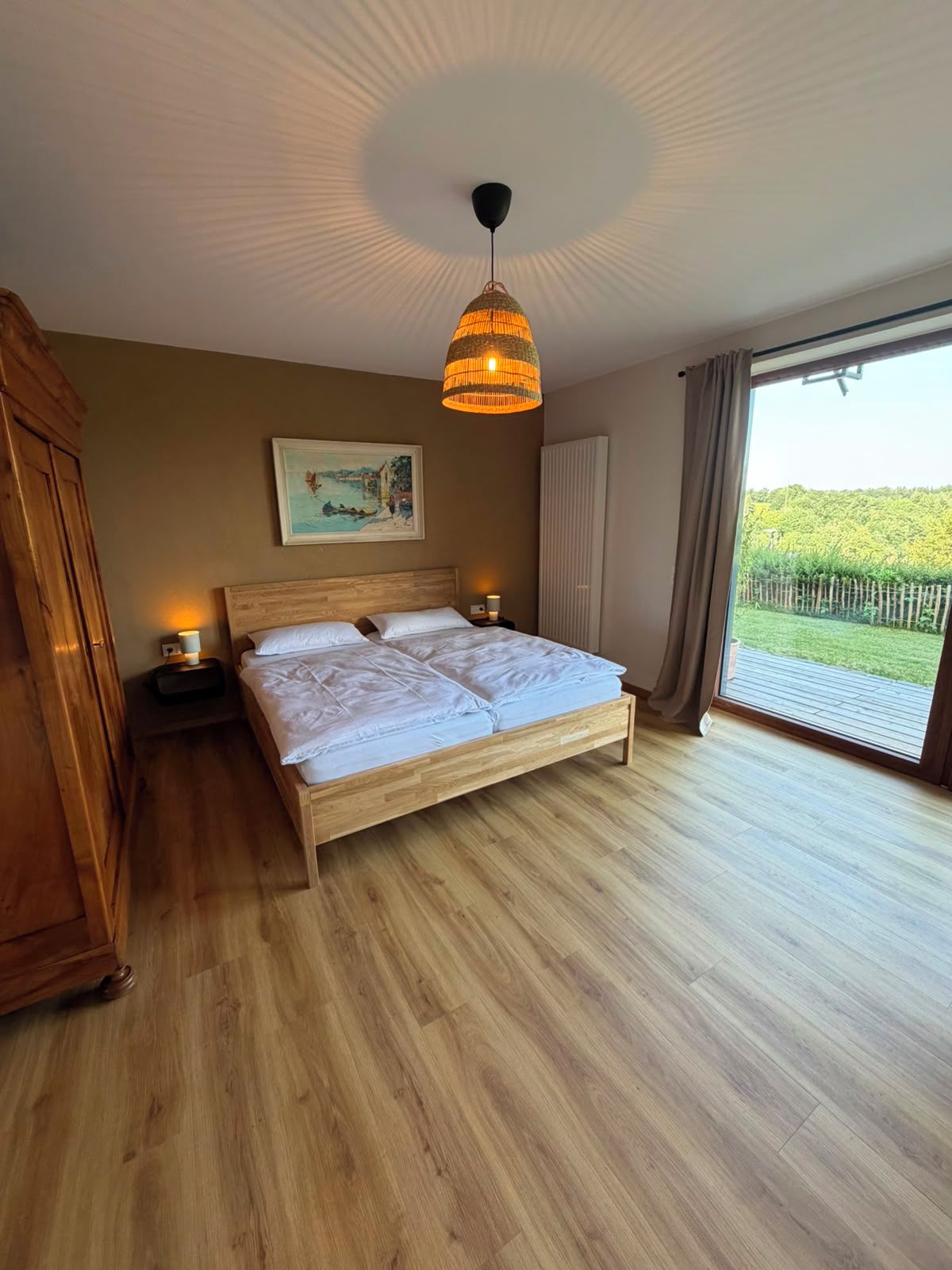 A bedroom with a wooden bed, two bedside lamps, a painting on the wall, and a large window with a view of greenery.