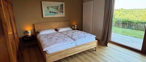 A bedroom with a wooden bed, two bedside lamps, a painting on the wall, and a large window with a view of greenery.