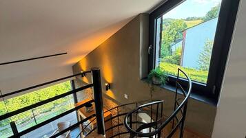 Un intérieur moderne avec un plancher en bois, un escalier en colimaçon et une grande fenêtre donnant sur un paysage verdoyant et un plan d’eau.