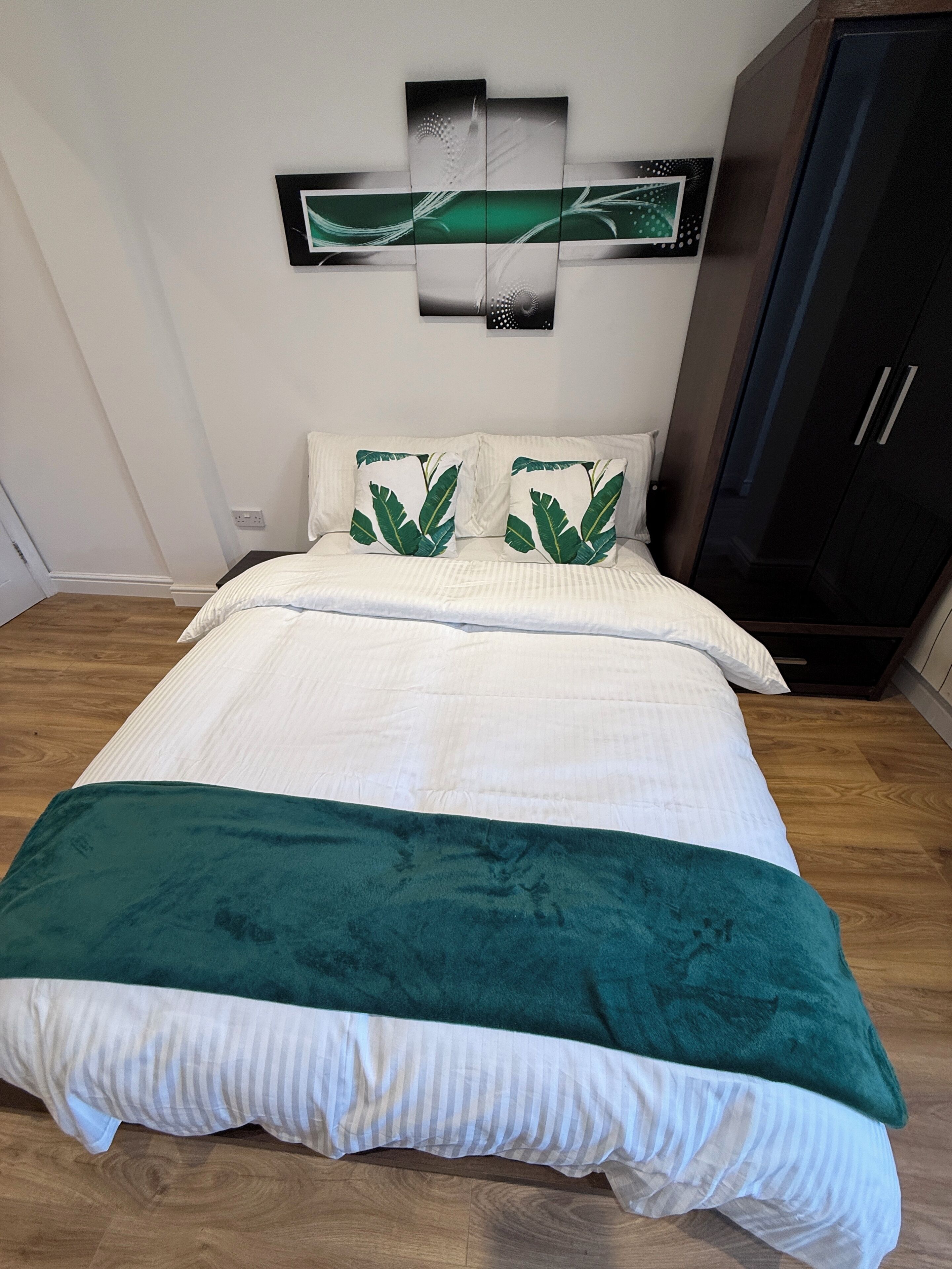 A neatly made bed with a white and green bedspread, two pillows with green leaf patterns, and a dark wooden wardrobe.