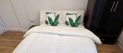 A neatly made bed with a white and green bedspread, two pillows with green leaf patterns, and a dark wooden wardrobe.
