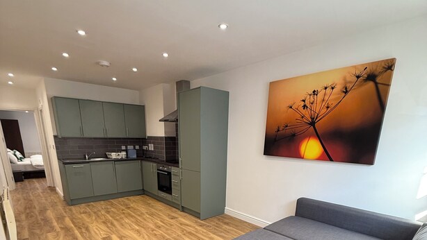 Un salón moderno con un sofá gris, piso de madera, una cocina al fondo y un cuadro de atardecer en la pared.
