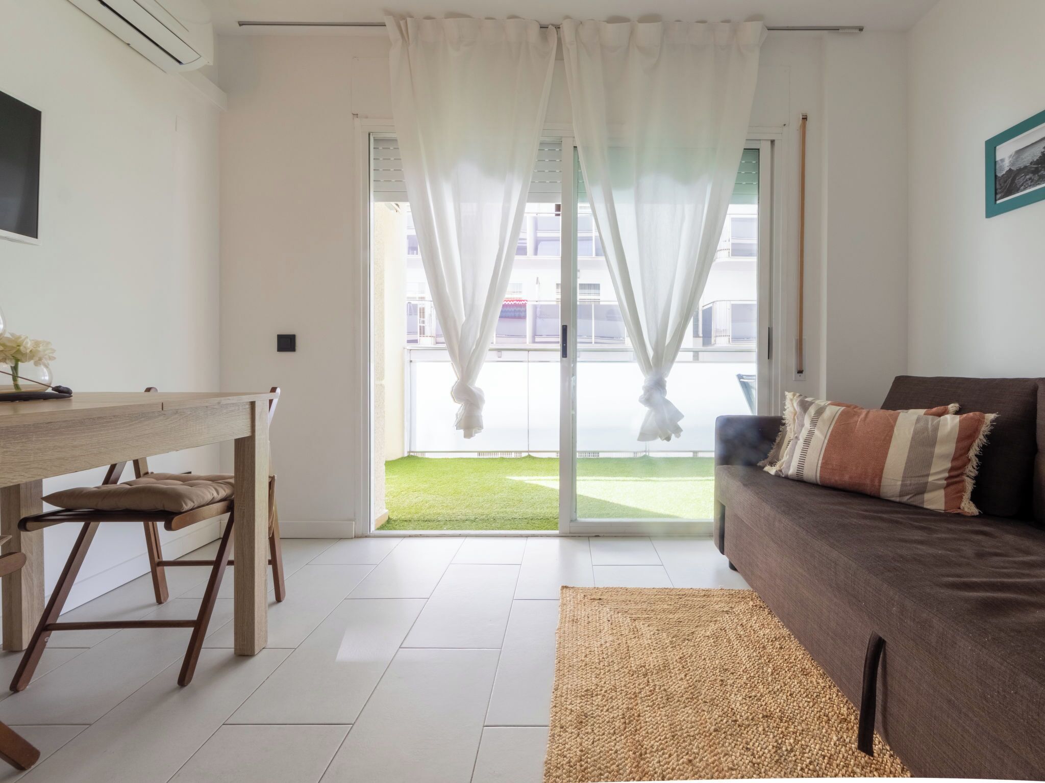 Un saló modern amb un sofà, una taula de dinar i vista a una zona exterior verda a través d'una porta de vidre.