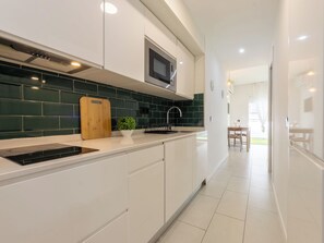 Uma cozinha moderna com armários brancos, uma placa de indução preta, uma tábua de corte de madeira, uma planta em vaso e um micro-ondas preto.