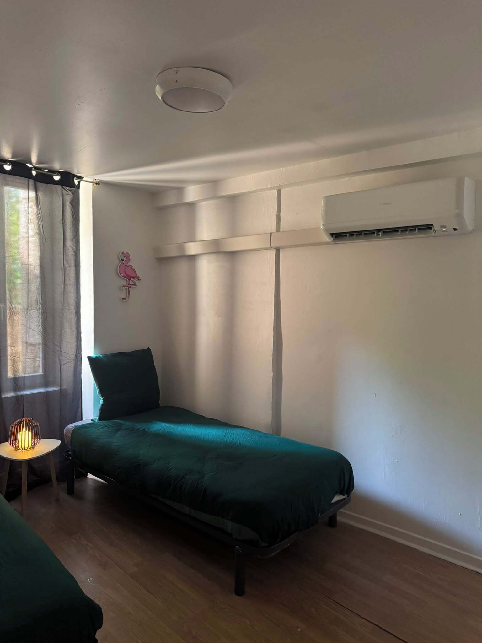 A single bed with a green cover, a small table with a lamp, a wall-mounted air conditioner, and a window with sheer curtains.