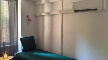 A single bed with a green cover, a small table with a lamp, a wall-mounted air conditioner, and a window with sheer curtains.