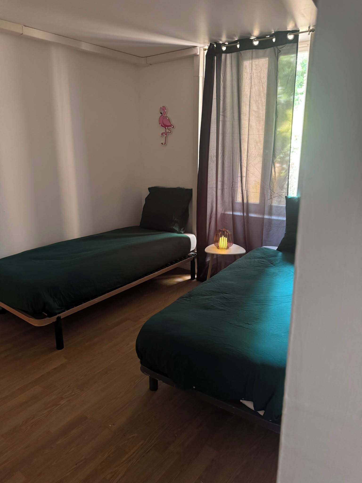 Een kamer met twee eenpersoonsbedden, een klein tafeltje met een lamp, een flamingo decoratie aan de muur en een raam met dunne gordijnen.