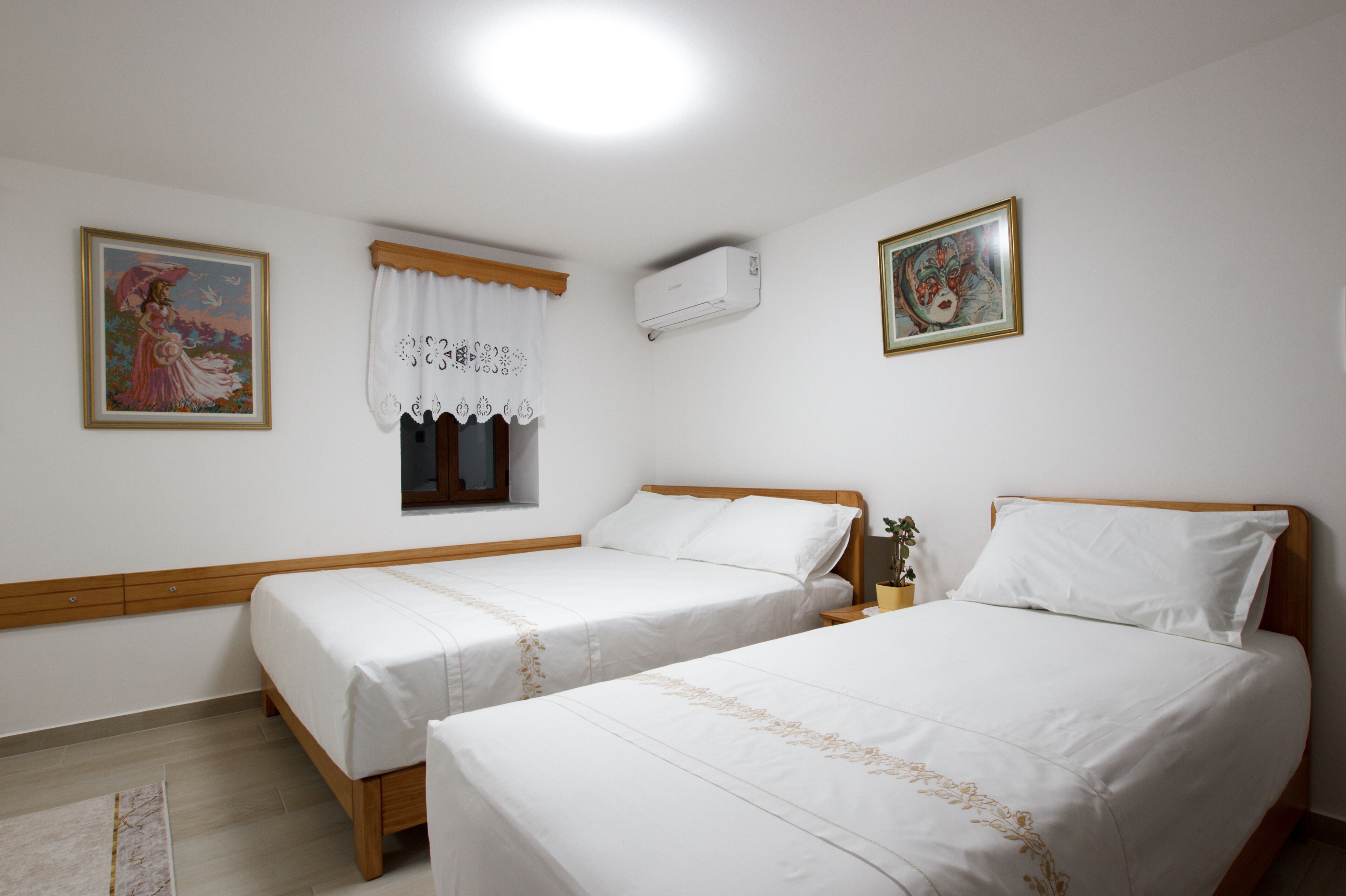 A room with two single beds, a window with a white curtain, an air conditioning unit, and two framed pictures on the walls.