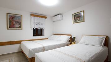 A room with two single beds, a window with a white curtain, an air conditioning unit, and two framed pictures on the walls.