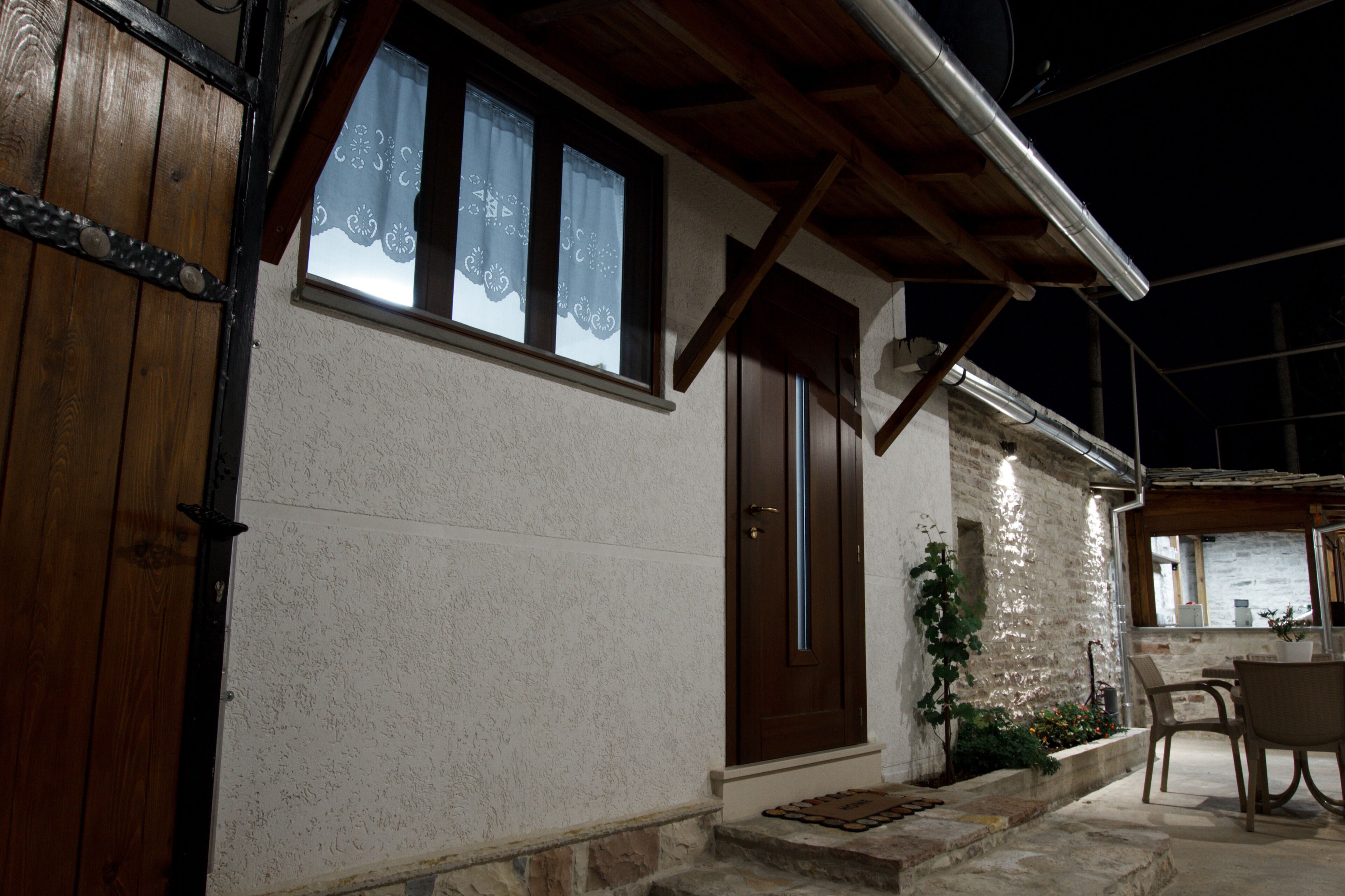 Un edificio con puerta de madera, ventana con cortinas de encaje, pared de piedra y un pequeño espacio al aire libre para sentarse.