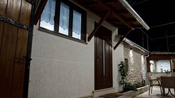 Un edificio con puerta de madera, ventana con cortinas de encaje, pared de piedra y un pequeño espacio al aire libre para sentarse.