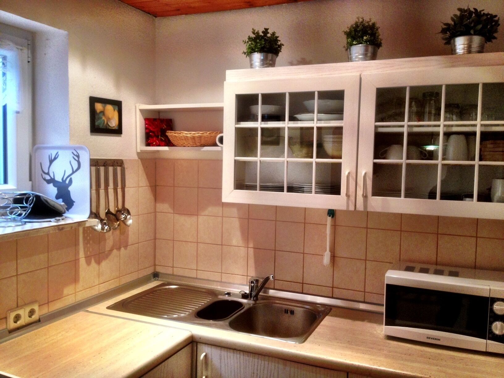A kitchen with a sink, a microwave, cabinets, and a window with a deer silhouette decoration.