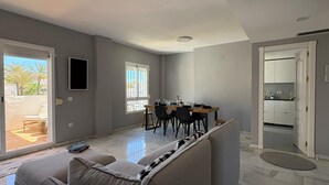 Ein modernes Wohnzimmer mit einer Couch, einem Essbereich mit Stühlen, einem an der Wand montierten Fernseher und einer durch eine offene Tür sichtbaren Küche.