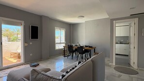 A modern living room with a sofa, a dining area with chairs, a TV mounted on the wall, and a kitchen visible through an open door. - Modern Duplex 3 Bedroom F72 (Marbella)