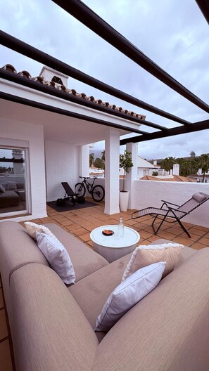A covered outdoor seating area with a sofa, a round table, and a bicycle. - Modern Duplex 3 Bedroom F72 (Marbella)