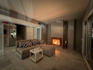 A modern living room with a sofa, ottoman, coffee table, fireplace, and a view of the kitchen. - Modern Duplex 3 Bedroom F72 (Marbella)