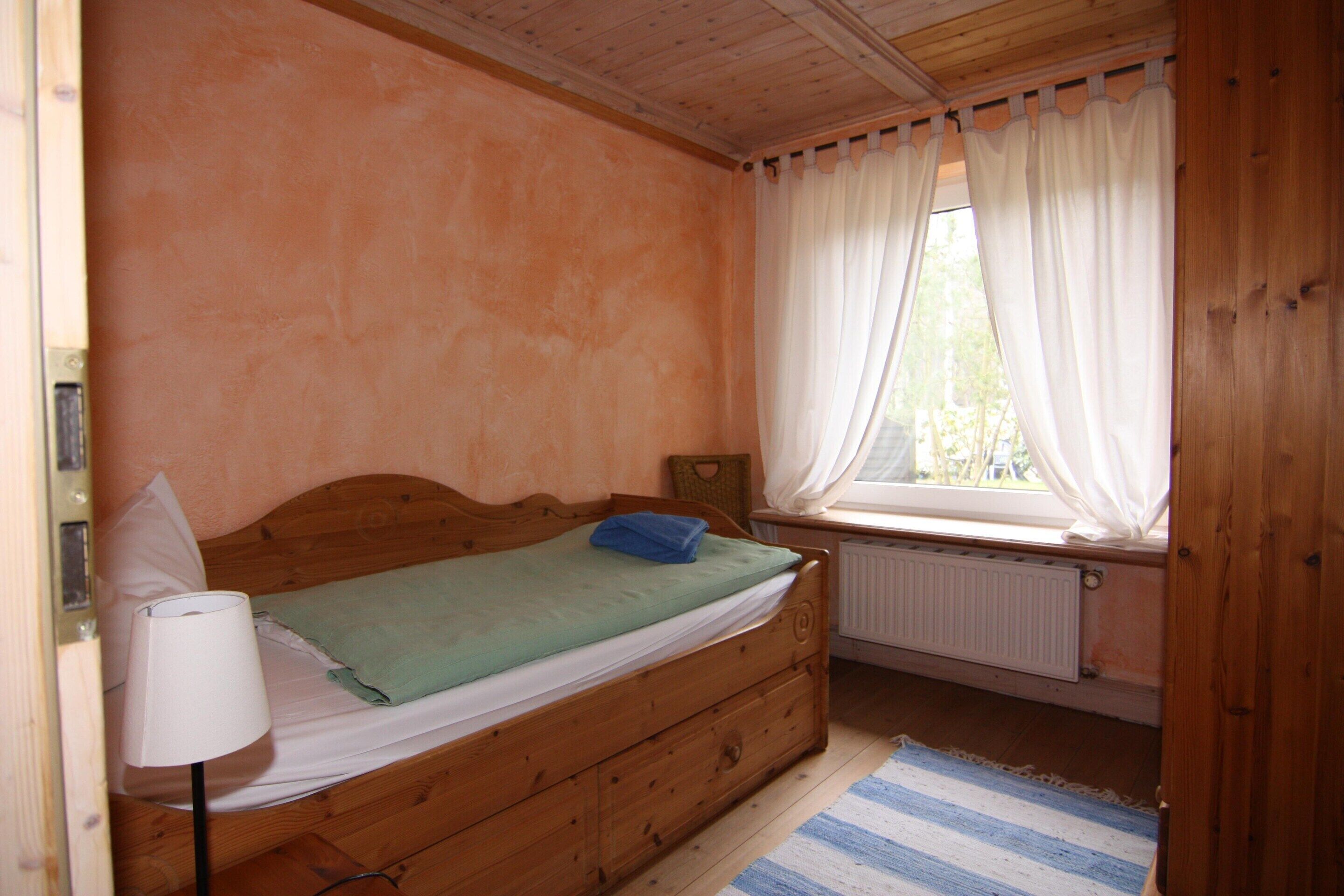 A single wooden bed with a green blanket, a white pillow, and a blue towel. There is a white lamp on the left side of the bed. A window with white curtains is visible, and a radiator is beneath it.