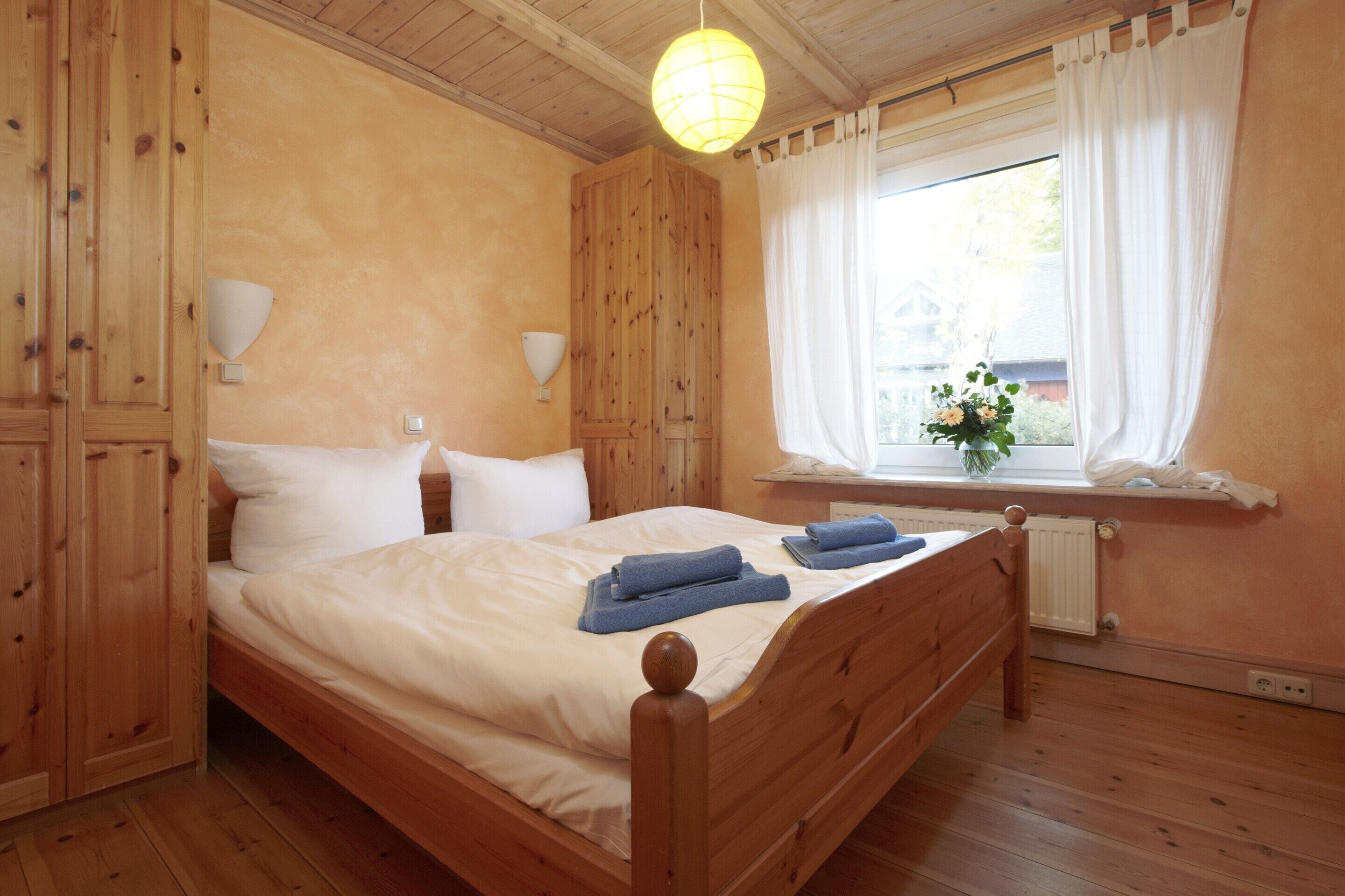 Un lit en bois avec du linge de lit blanc et des serviettes bleues, une armoire en bois, une table de chevet avec une lampe et une fenêtre avec des rideaux.