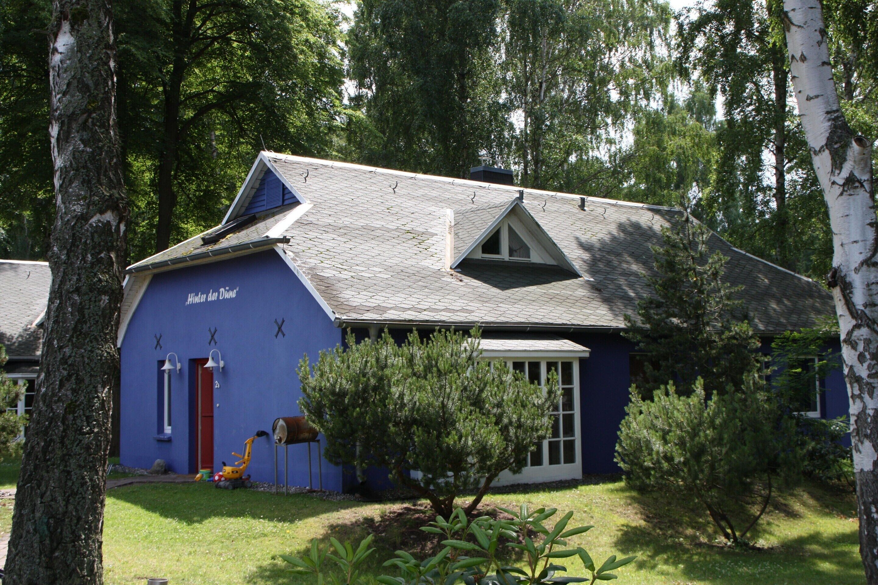 A house with a blue exterior, a red door, and a small garden with a tree and shrubs.