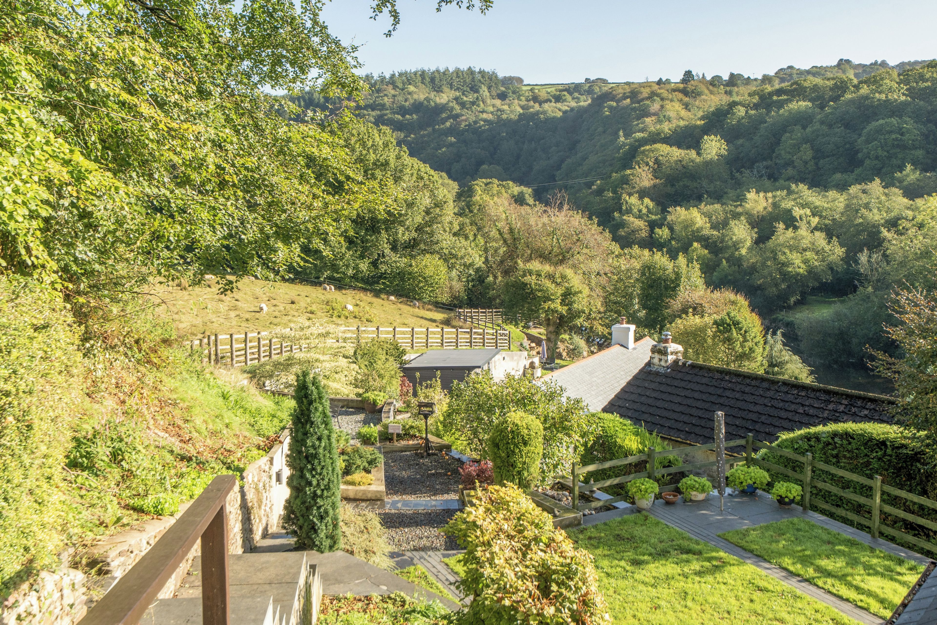 A hillside property with a stone pathway leading to a wooden deck, surrounded by lush greenery and a view of rolling hills.